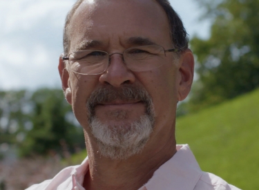 Headshot of Dr. Chudacoff: A white man with a grey beard and glasses, in a pink button down shirt, standing outside