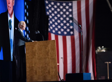 President Joe Biden giving a speech at a podium, with a large monitor and American flag in the background. Photo by Adam Schultz.