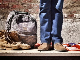 Photo by Benjamin Faust. The legs and boots of a veteran, standing next to a pair of boots, a camo backpack, and an American flag.