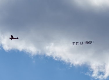 A plane flies a "STAY AT HOME" banner across a cloudy sky