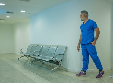 A man with a white beard wearing blue scrubs stands next to empty seats in a clinic hallway. Photo by Foto Garage AG on Unsplash