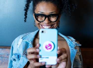 A young black woman wearing thick black glasses and a denim jacket smiles into her smartphone cameria
