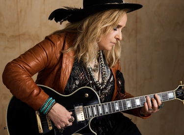 Melissa Etheridge plays guitar in a brown leather jacket and wide-brimmed black hat
