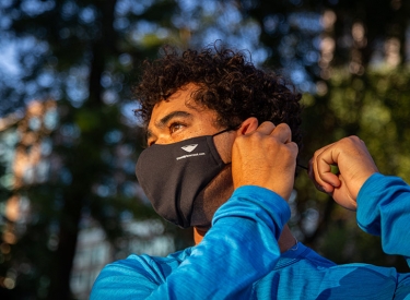 A young man with curly hair in a blue long sleeved tee puts on his face mask