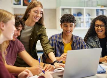 A group of students working together, focused on their laptops and taking notes in a classroom setting.